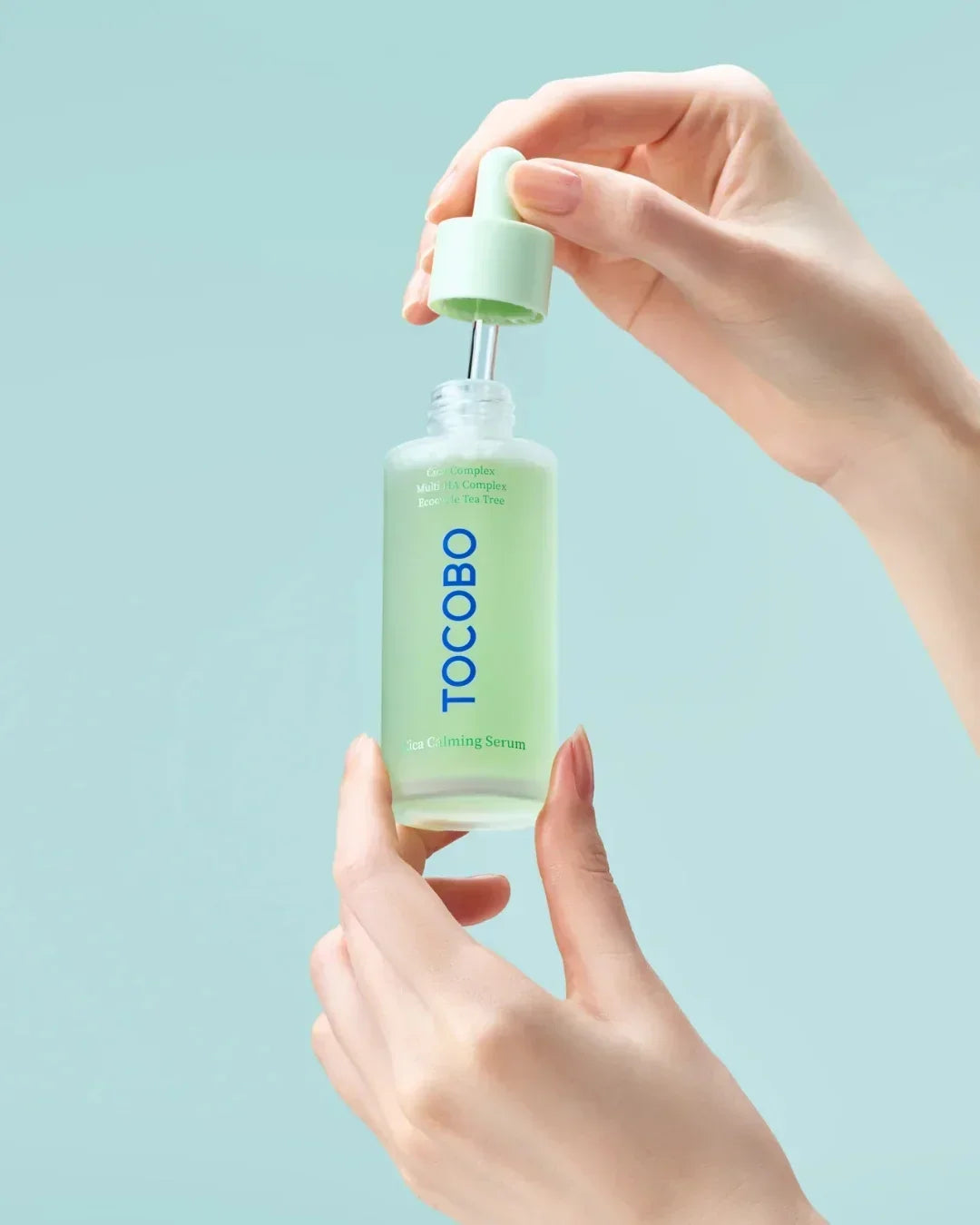Hands holding and opening TOCOBO Cica Calming Serum bottle with dropper on light blue background