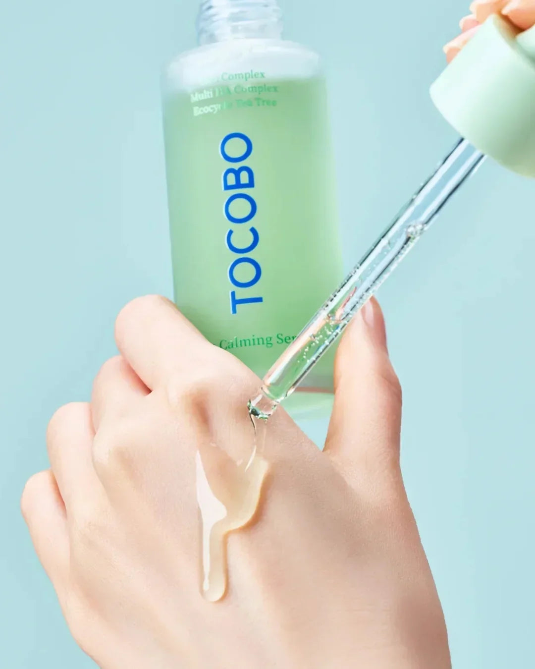 Hand applying clear serum drop from Tocobo calming serum bottle with dropper on light blue background