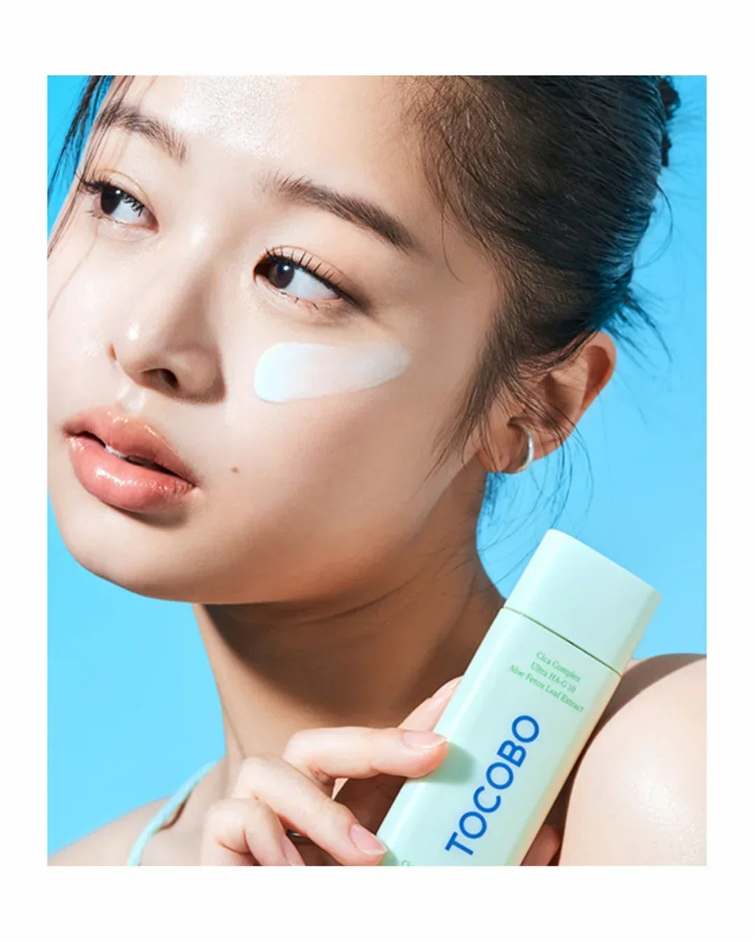 Close-up of woman with white cream on cheek holding TOCOBO skincare serum bottle against blue background
