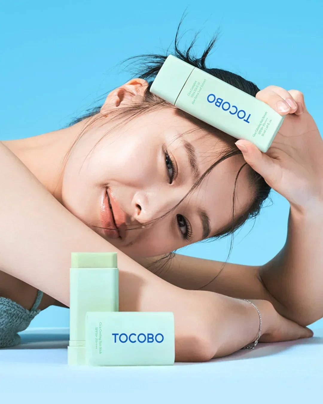 Smiling woman with clear skin holding green TOCOBO sunscreen stick against blue background