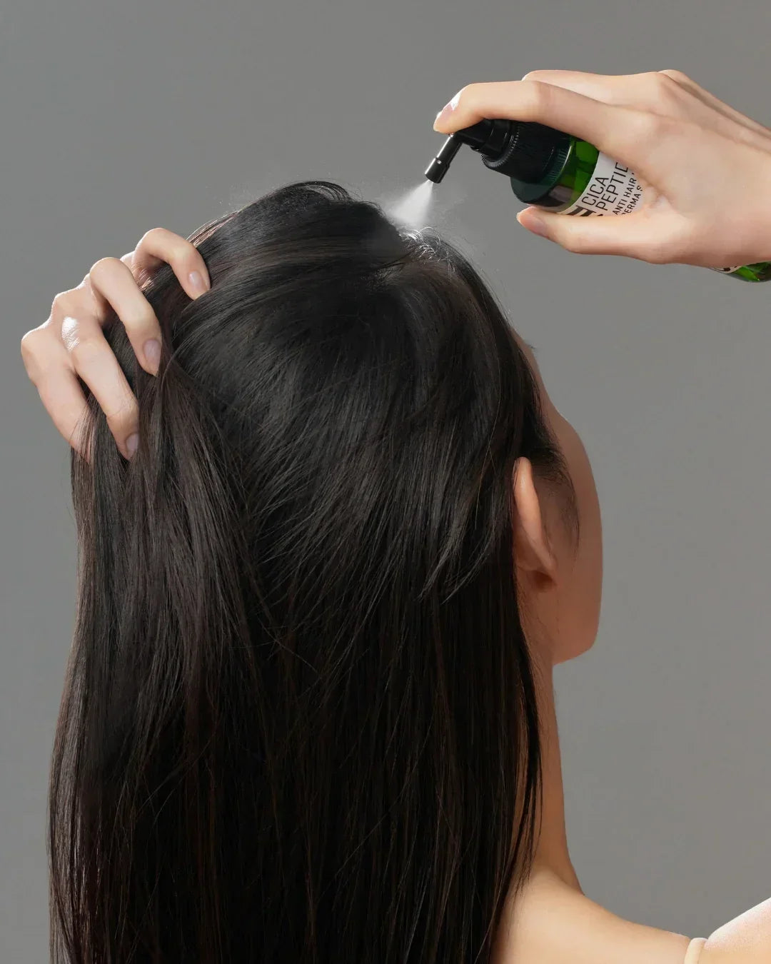 Woman spraying Cica Peptide anti-hair loss scalp tonic onto dark hair against gray background