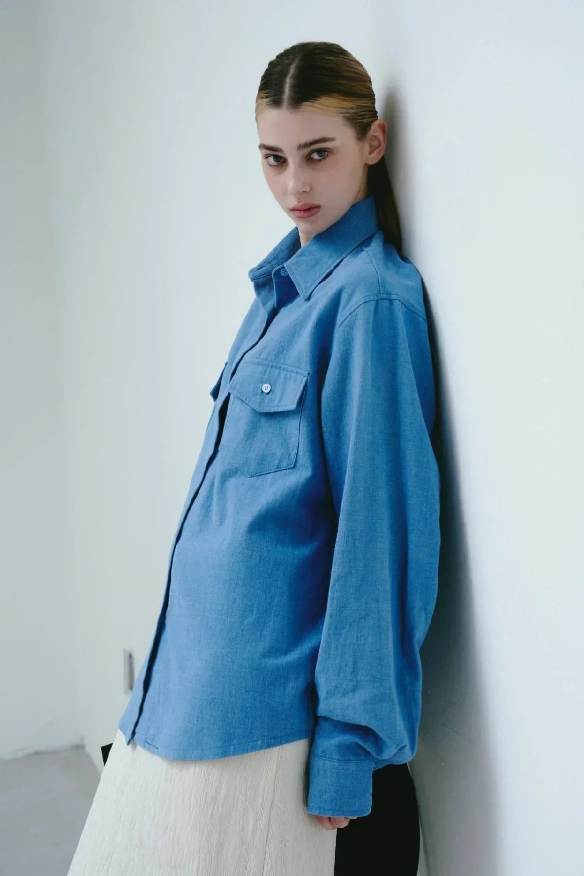Young woman wearing light blue classic denim overfit shirt leaning against white wall