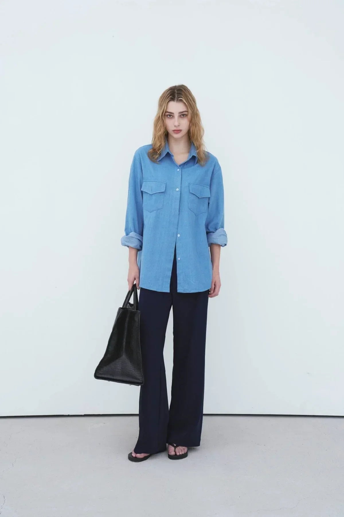 Model wearing light blue oversized denim shirt with rolled sleeves, black wide-leg pants, and holding black leather tote bag