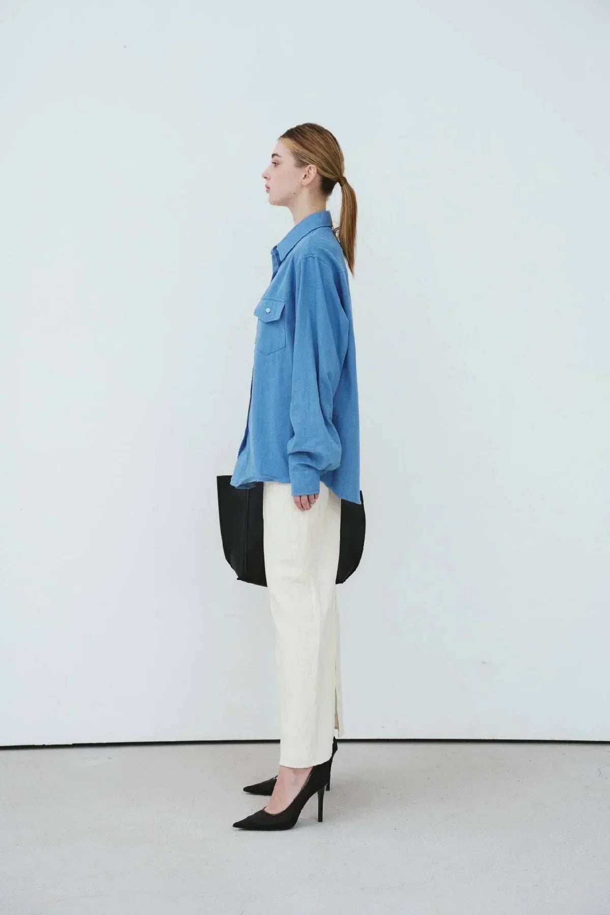 Side view of woman wearing light blue oversized denim shirt, white pants, black heels, and carrying black bag