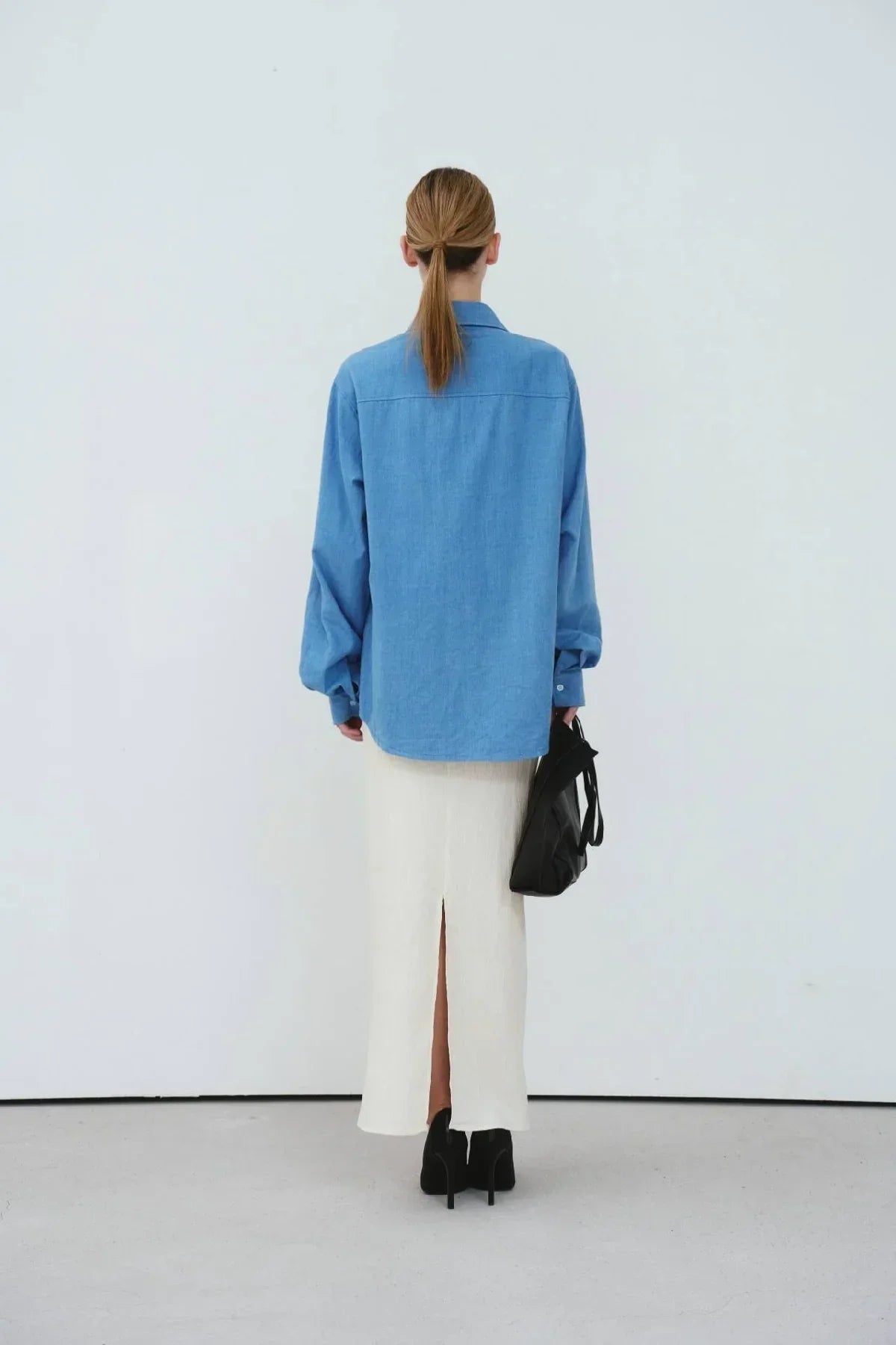 Woman wearing light blue oversized denim shirt with white midi skirt and black heels, holding black bag