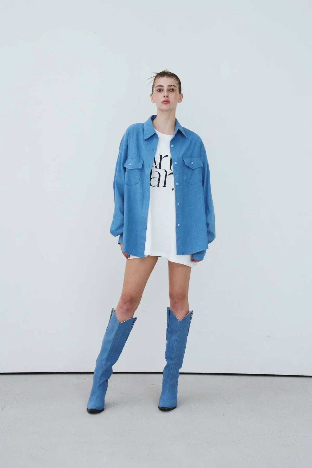 Model wearing light blue oversized denim shirt and matching denim knee-high boots in a studio
