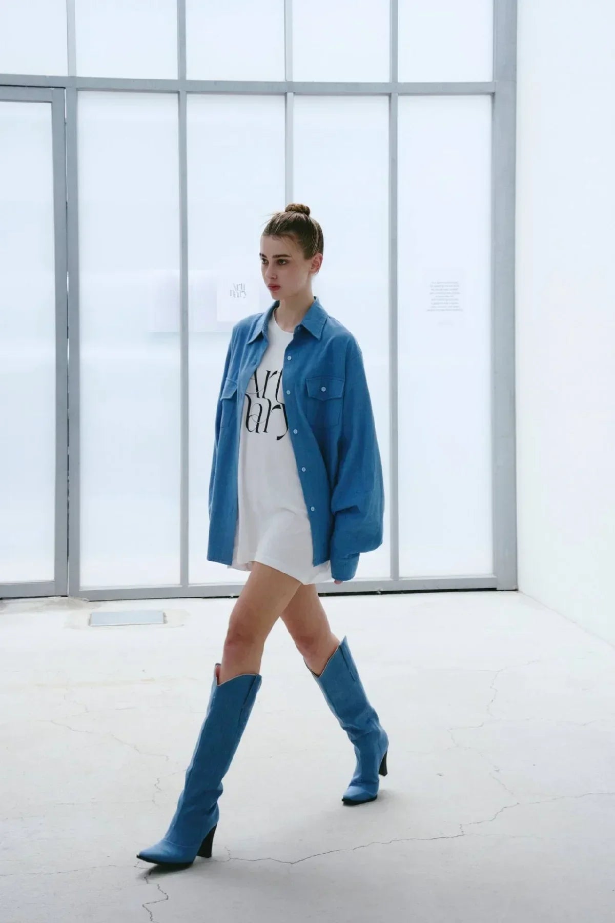 Woman wearing light blue oversized denim shirt and matching boots with white dress in bright minimal studio