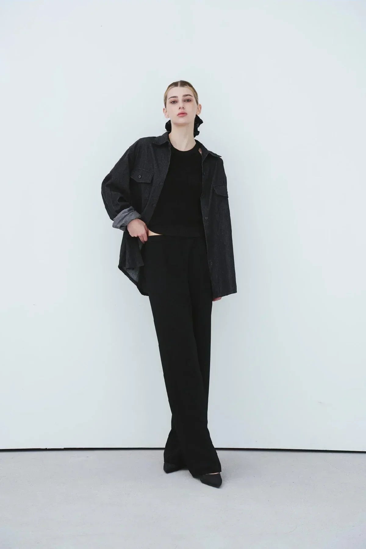 Woman wearing black ribbed top, oversized dark shirt, wide-leg black pants, and black heels against white wall