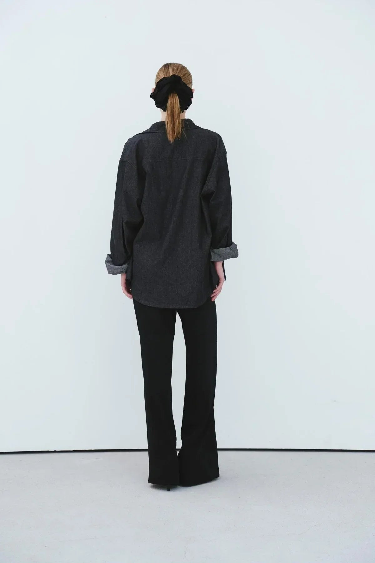 Woman with ponytail wearing black oversized denim shirt and black pants, standing against white wall