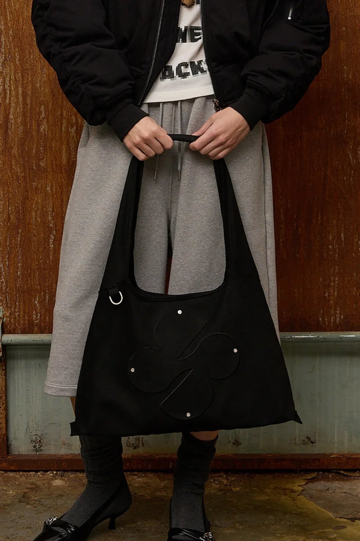 Person holding black artificial suede shoulder bag with clover design, dressed in black jacket, grey skirt, and black heels