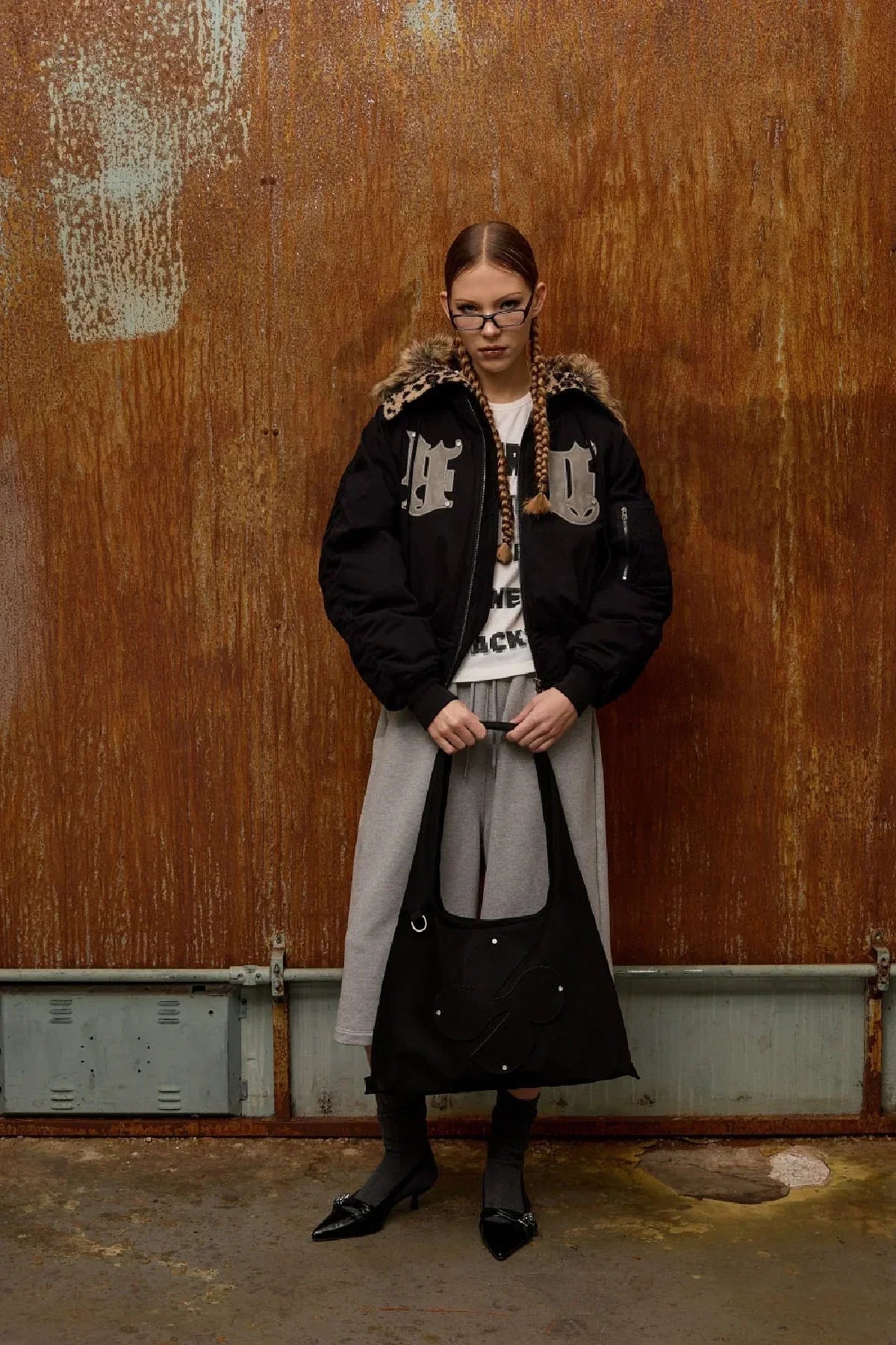 Model wearing black bomber jacket with fur collar, gray skirt, holding black suede shoulder bag with clover logo