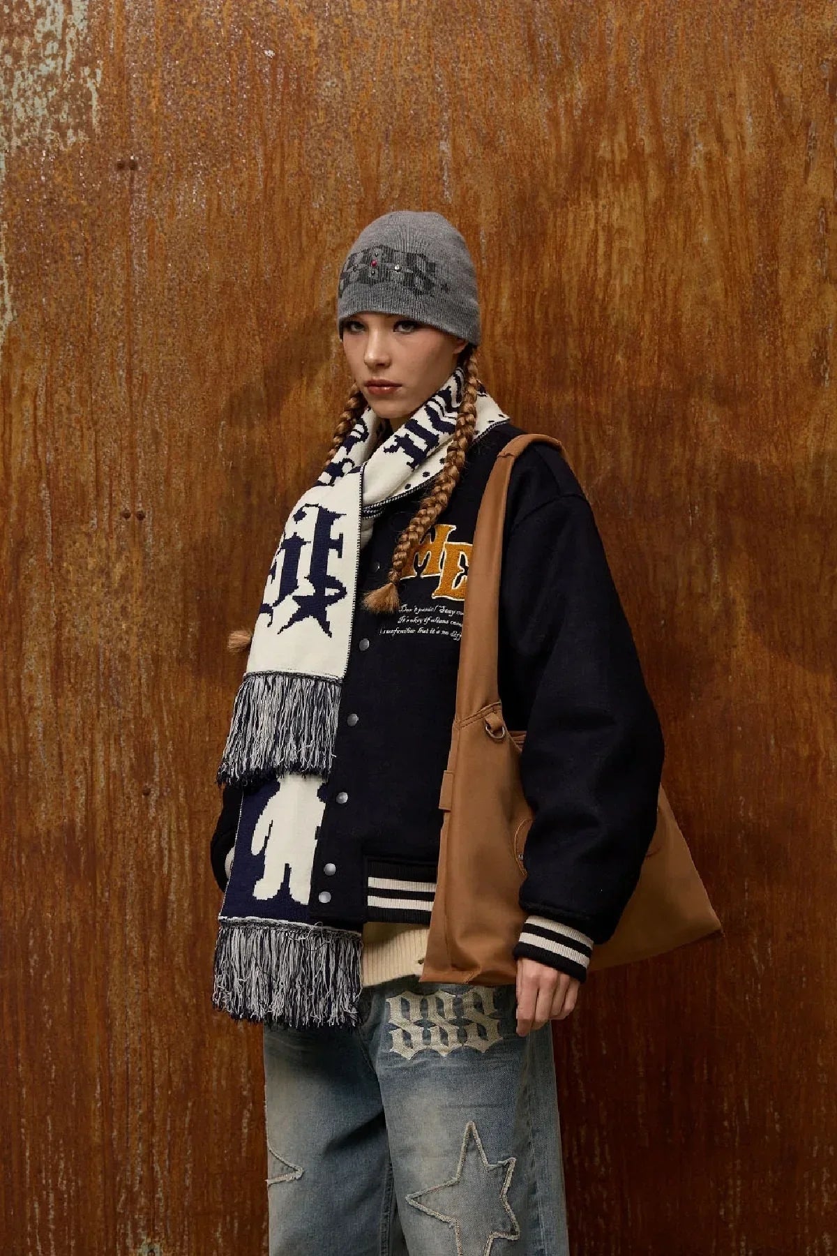Young woman in gray beanie, black varsity jacket, patterned scarf, denim jeans with star patches, and brown shoulder bag