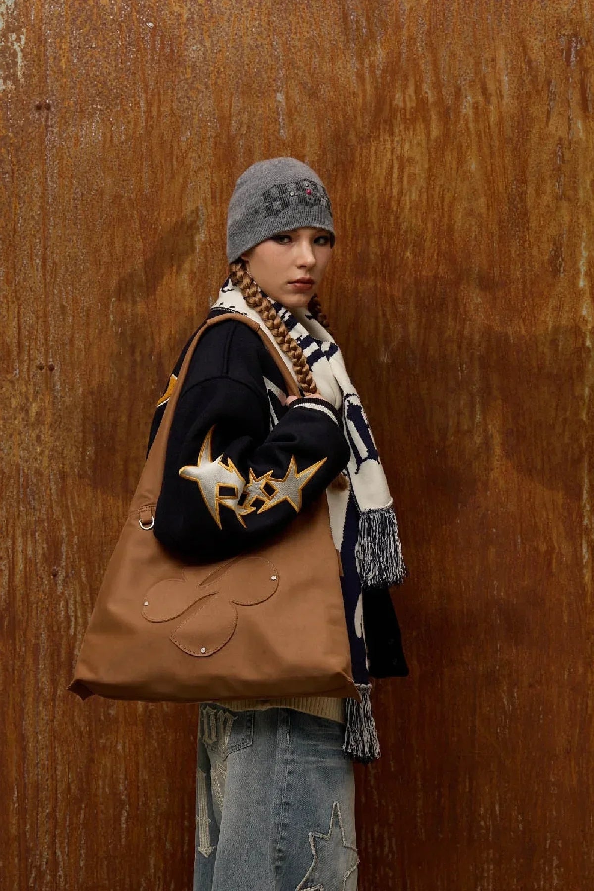 Model wearing grey beanie, black jacket with star patches, scarf, and carrying camel artificial suede shoulder bag