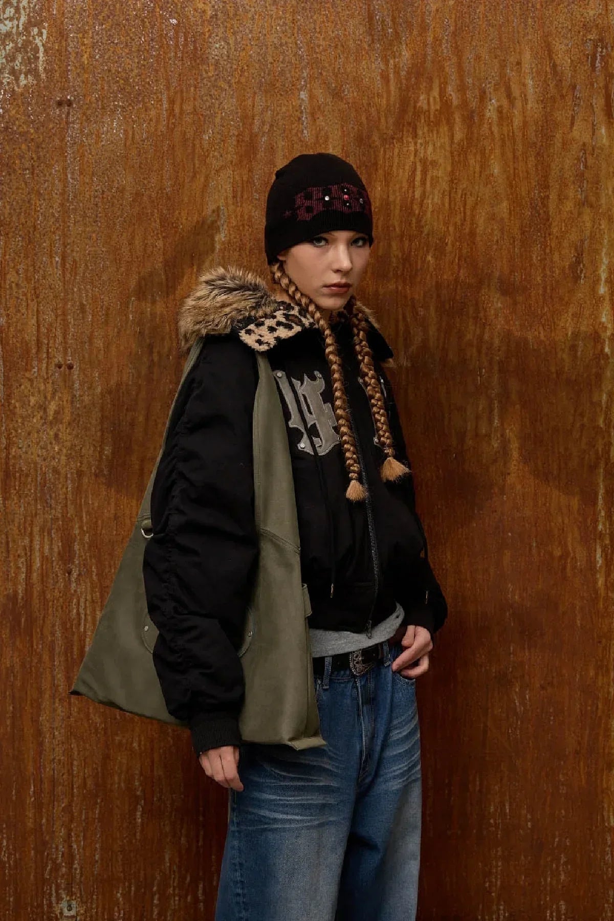 Model in black fur-collared jacket, black beanie, blue jeans, and khaki artificial suede shoulder bag