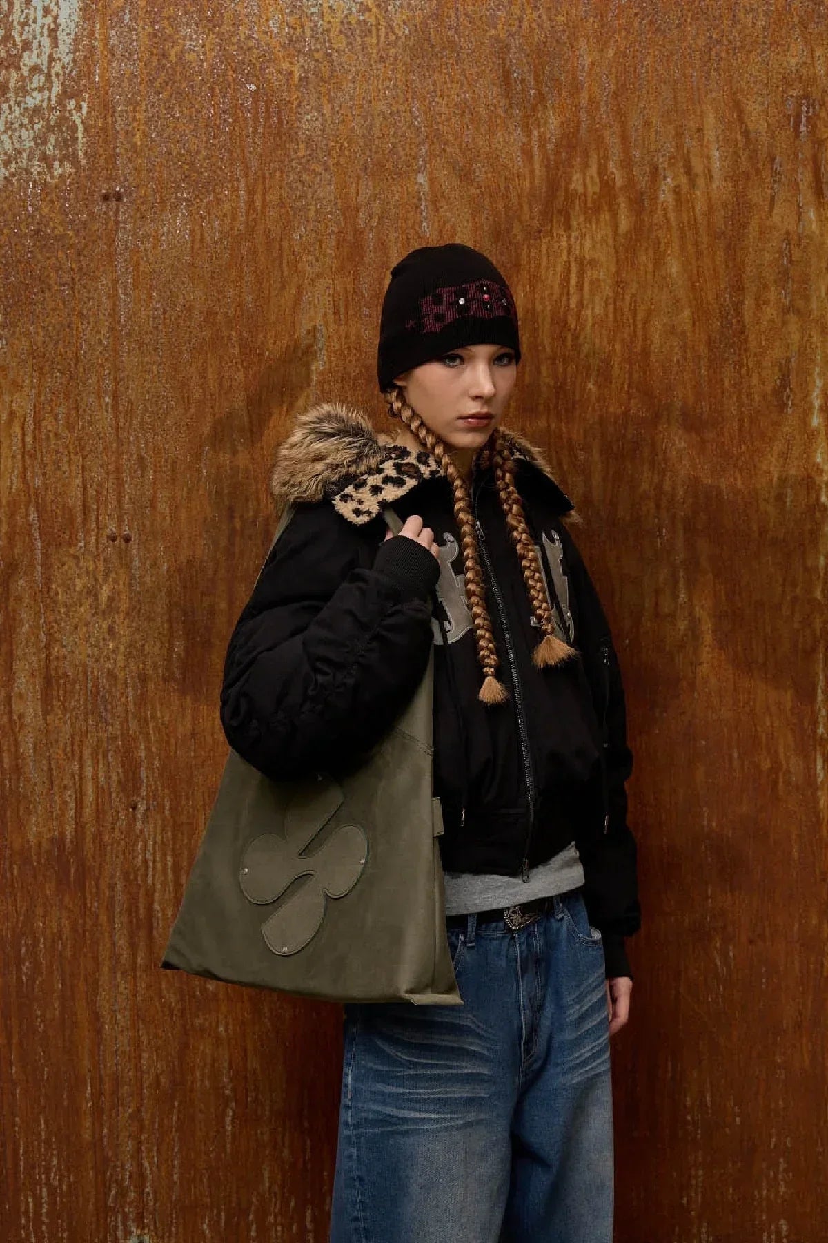 Young woman with two braids wearing black beanie, fur-collared jacket, blue jeans, and khaki artificial suede shoulder bag