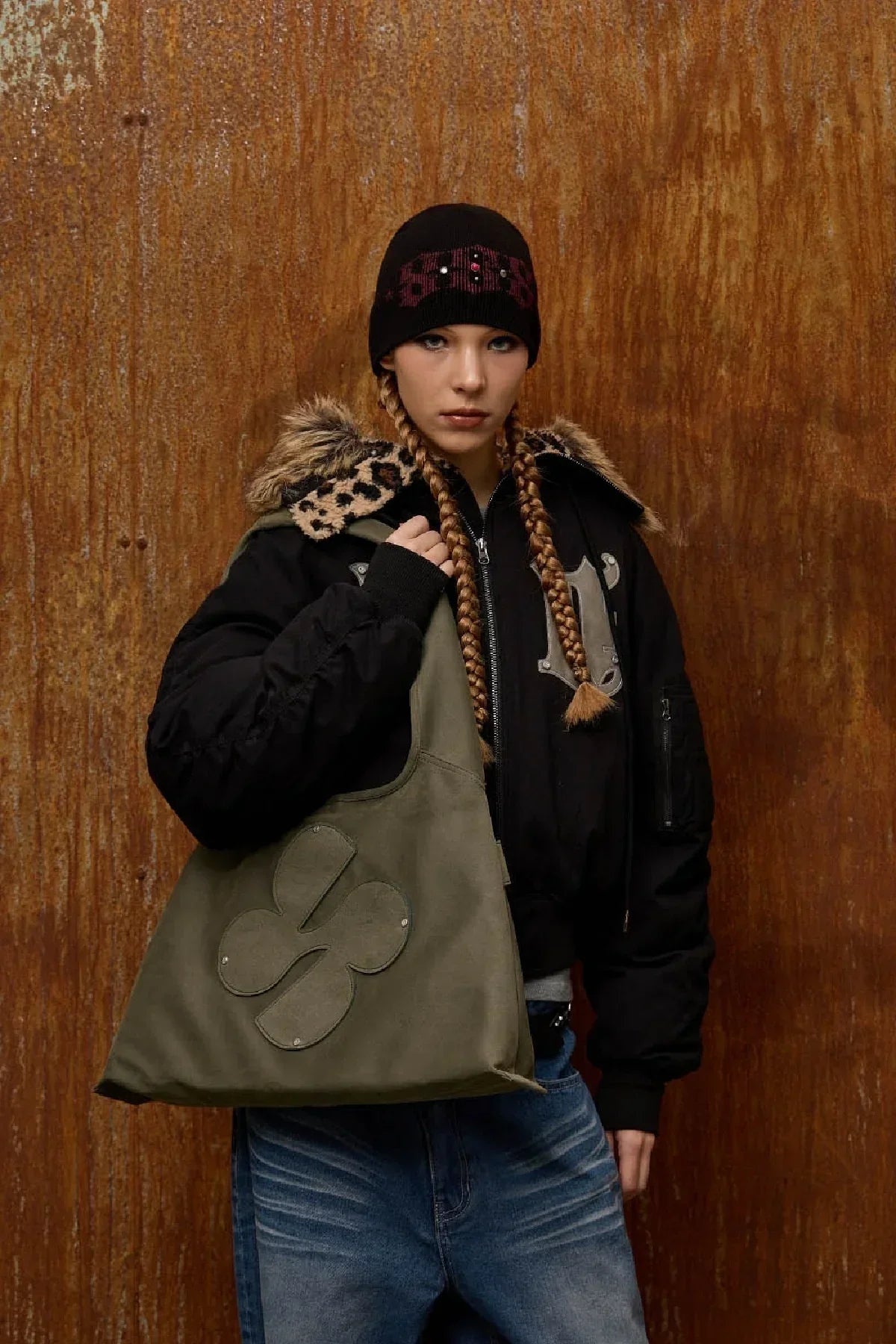 Young woman with braided hair wearing black jacket and beanie, holding khaki artificial suede shoulder bag with clover logo