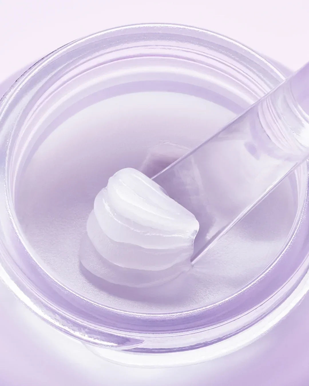 Close-up of translucent collagen-boosting lip mask cream texture with spatula in clear jar on soft purple background