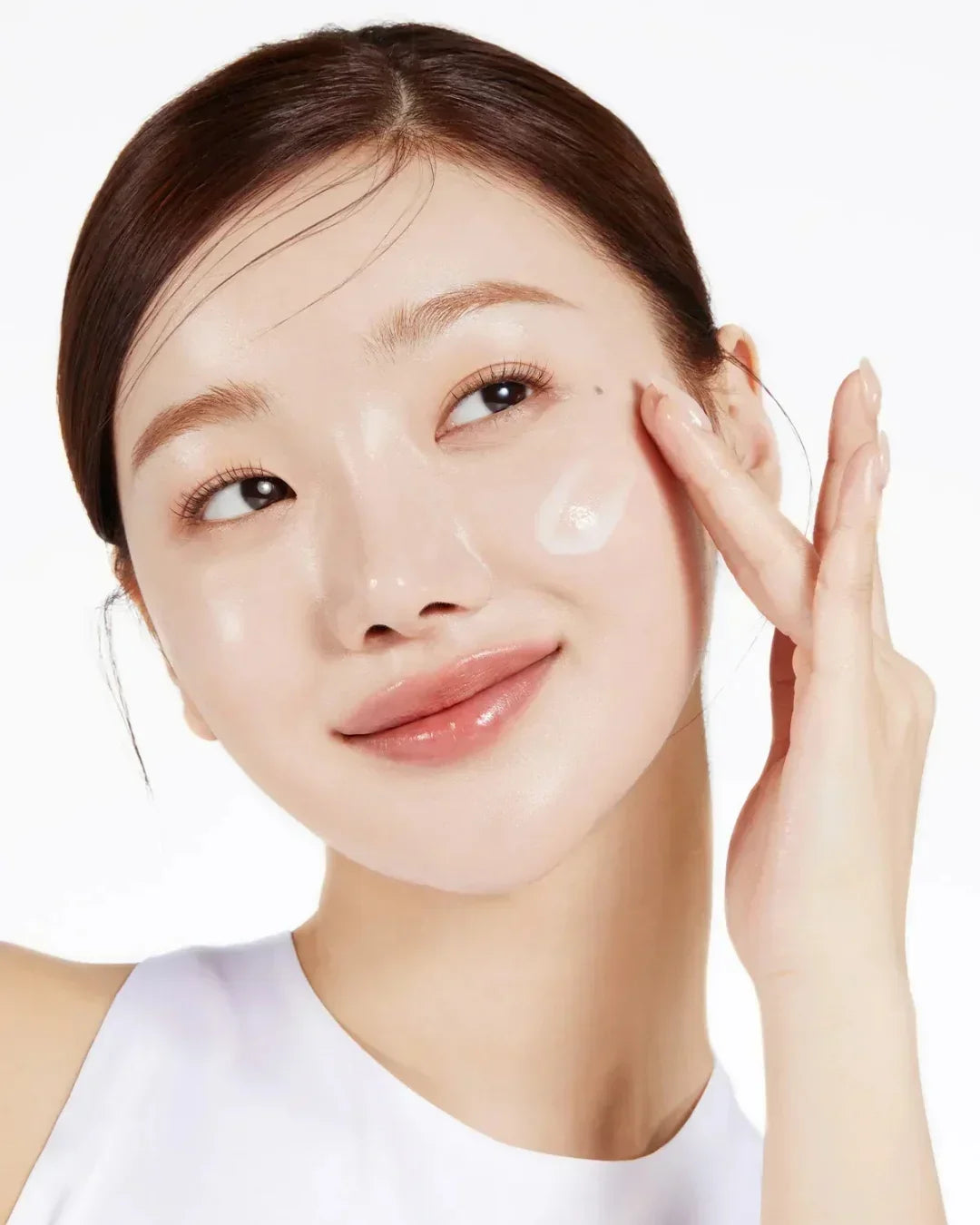 Close-up of a woman with clear skin applying white cream to her cheek, glowing skincare routine
