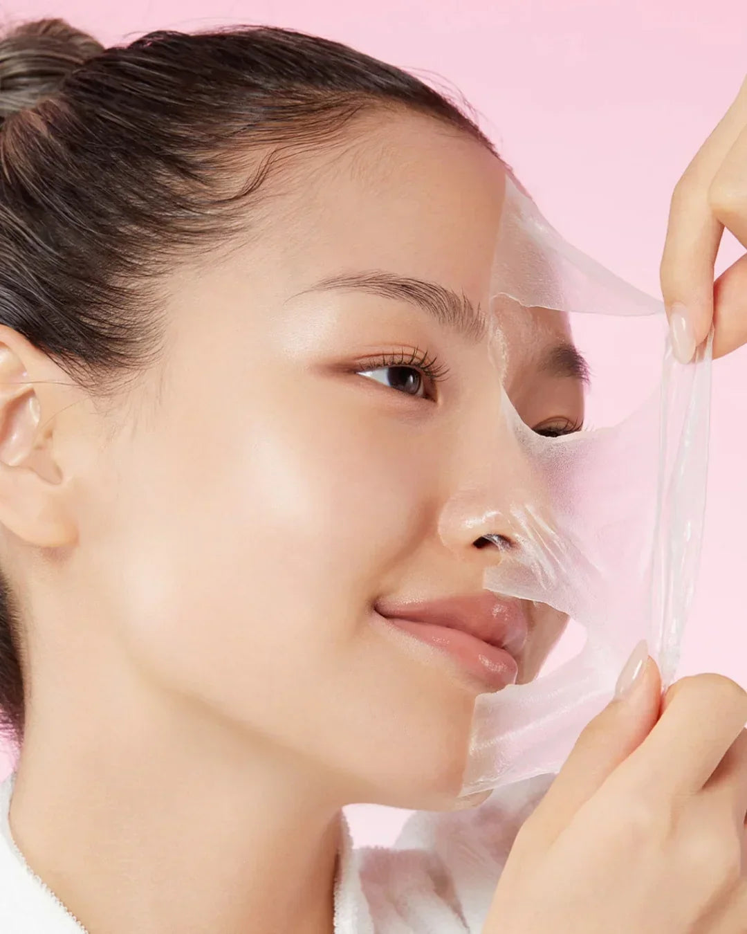 Young woman peeling off clear collagen night mask from smooth glowing skin on pink background