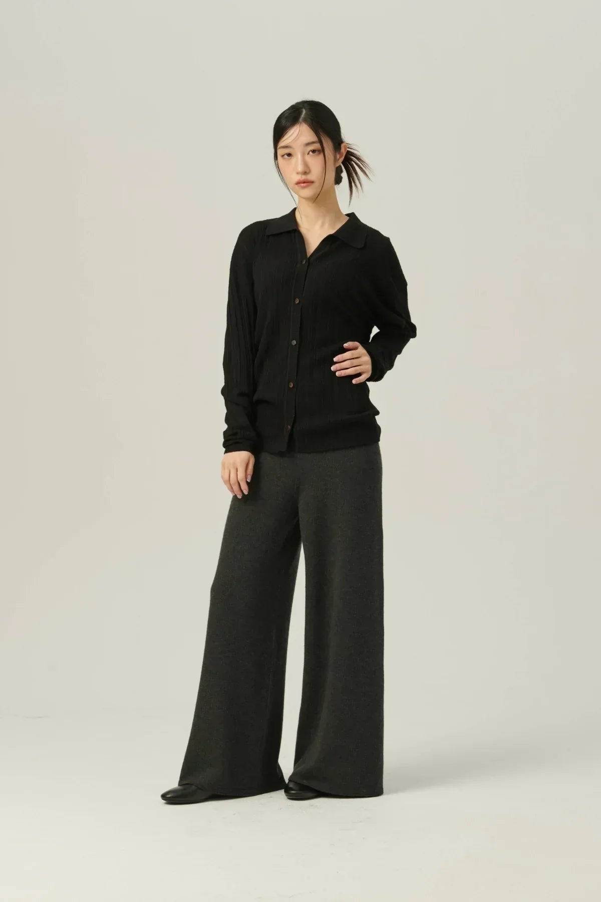 Woman wearing black collared rib knit cardigan and wide-leg dark gray pants on plain background