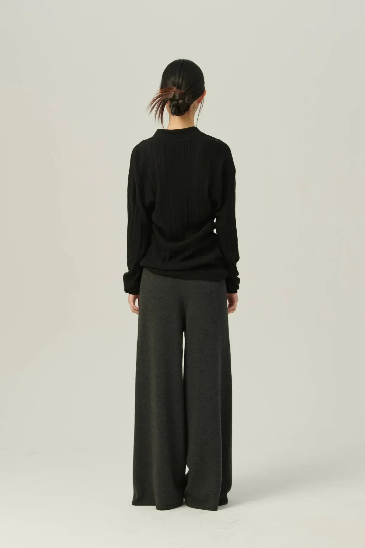 Woman wearing black collared rib knit cardigan and wide-leg gray pants, standing against a plain background