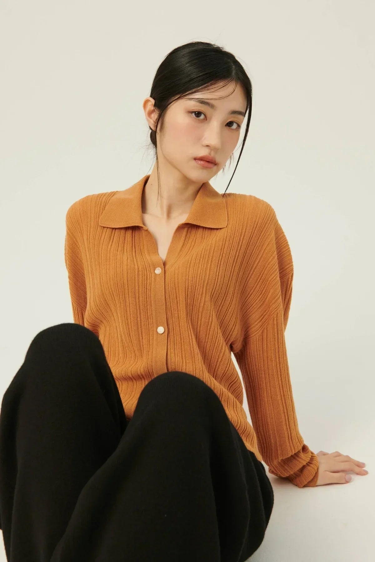 Young woman wearing camel rib knit collared cardigan with buttons, paired with black pants, seated pose