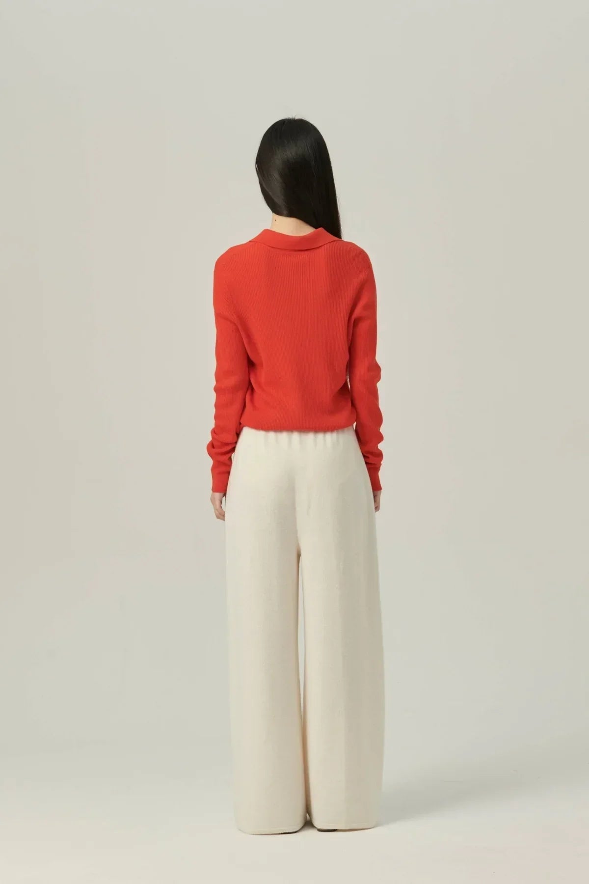 Back view of woman wearing red collared rib-knit cardigan and wide cream knit pants on plain background
