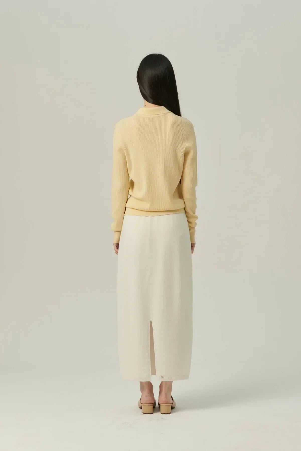 Woman wearing a yellow collared rib-knit cardigan and white midi skirt with back slit, standing on plain background