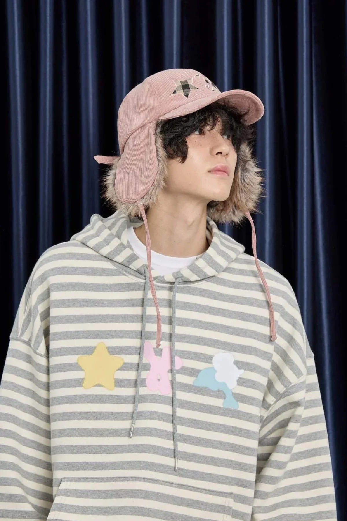 Young man wearing a pink corduroy trooper cap with faux fur lining and a gray striped hoodie with colorful applique