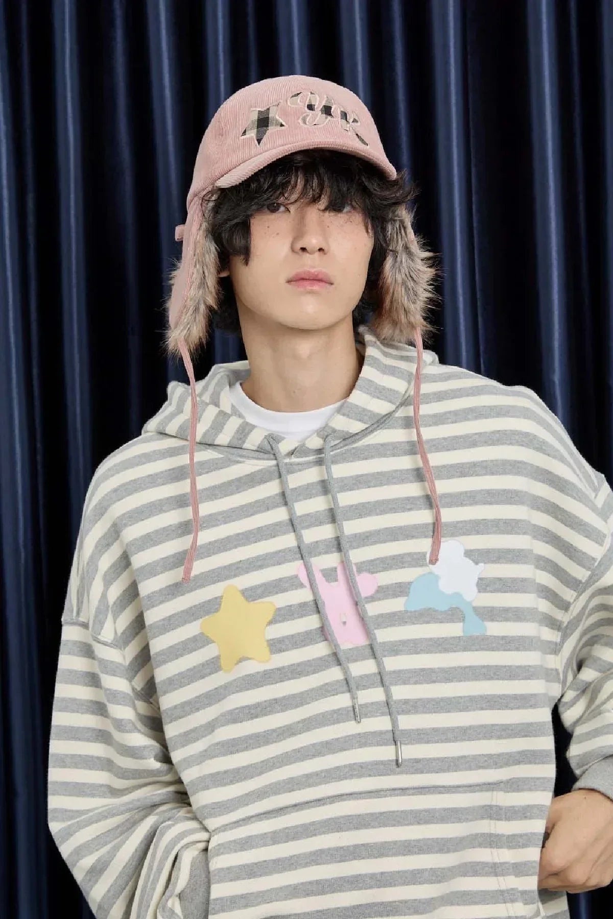 Man wearing pink corduroy trooper cap with plaid applique and striped hoodie with star and animal patches