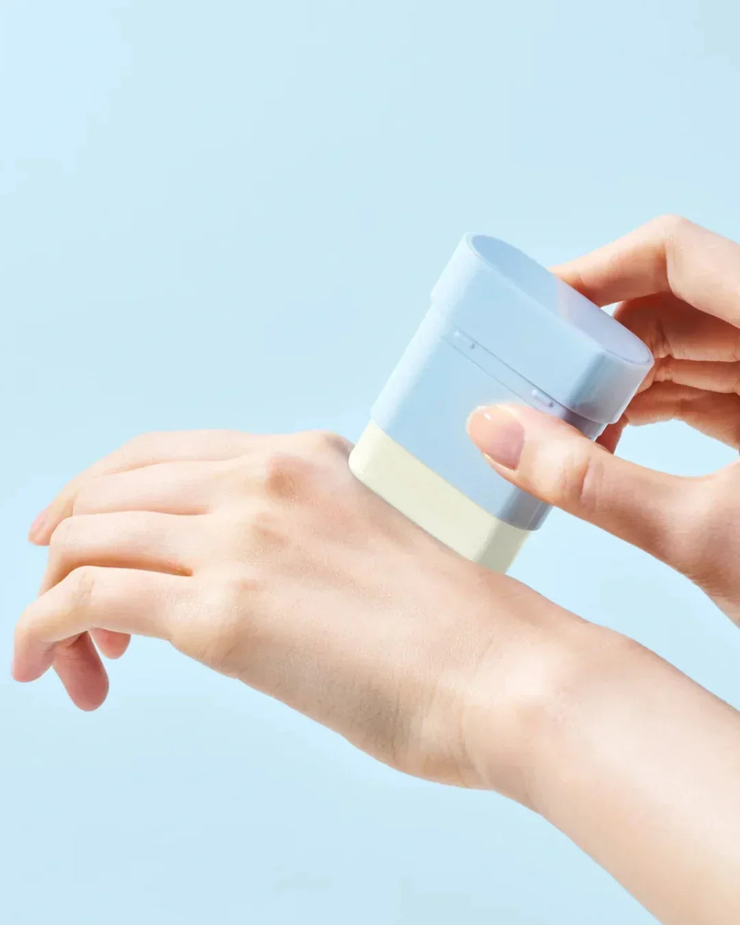 Hand applying blue and white sun stick sunscreen on back of hand against light blue background