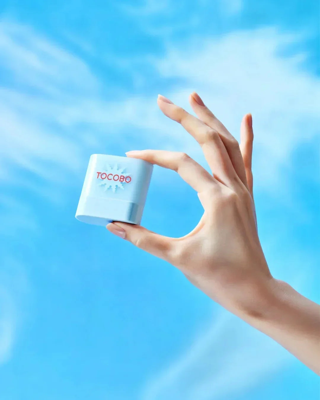 Hand holding small white TOCOBO skincare product against bright blue sky background