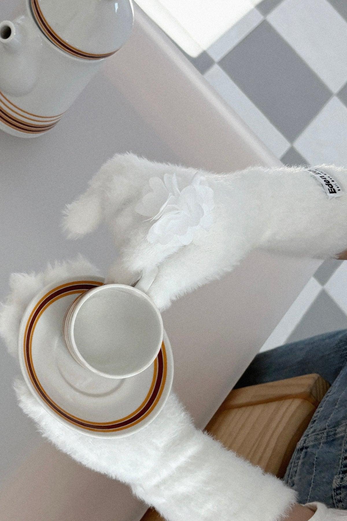 Hands wearing white fuzzy ring gloves with flower applique holding a white cup and saucer indoors
