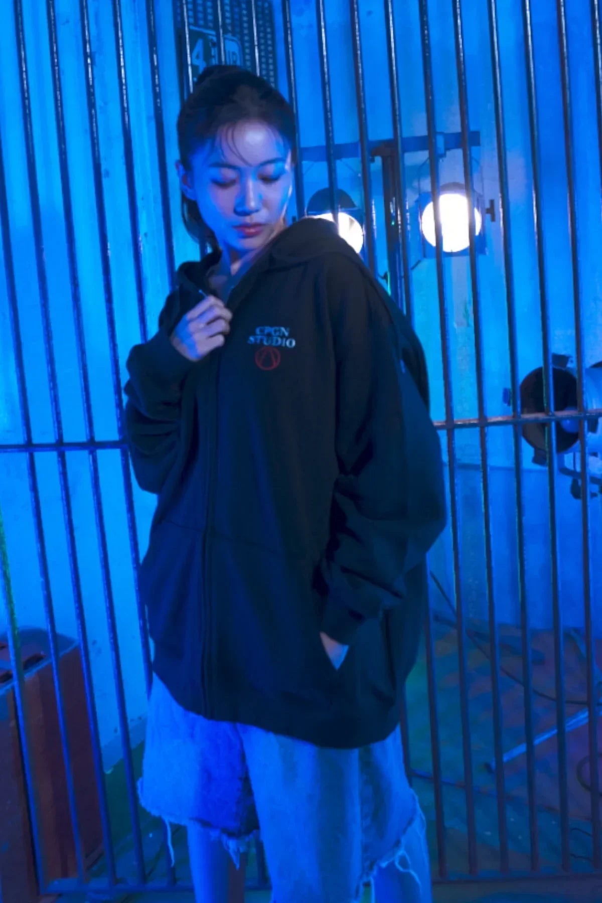 Young woman wearing black CPGN STUDIO zip-up hoodie and denim shorts in blue lit industrial setting with metal bars