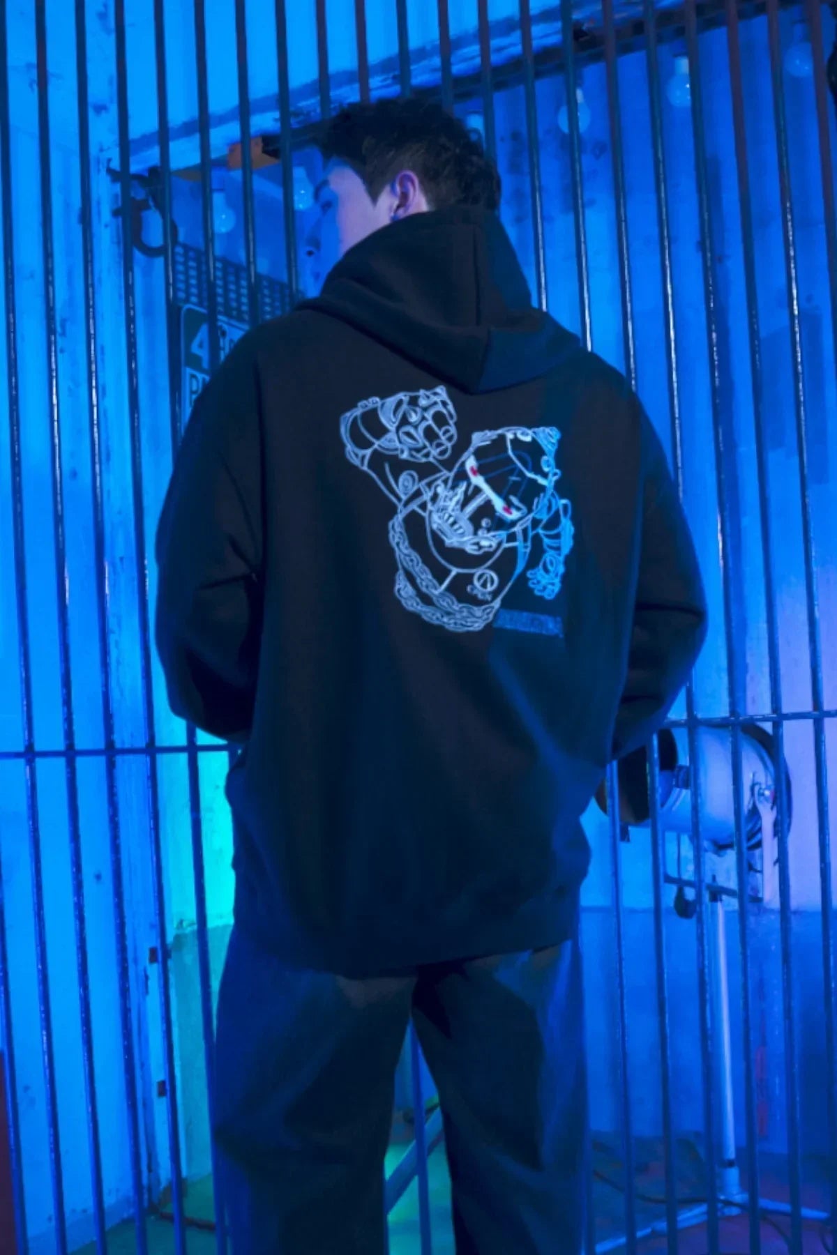 Man wearing black hoodie with robotic fist graphic, standing in blue-lit metal gate setting