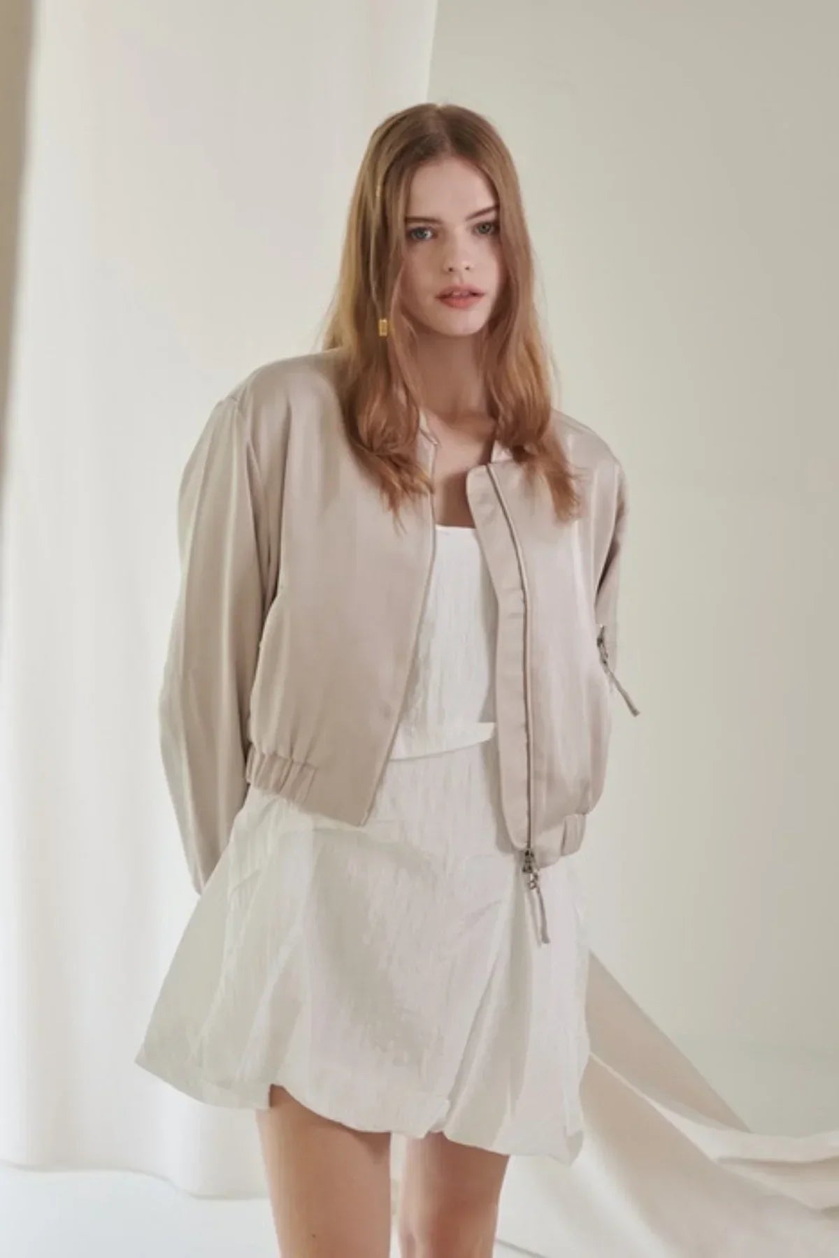 Young woman wearing beige satin bomber jacket over white dress in minimal studio setting