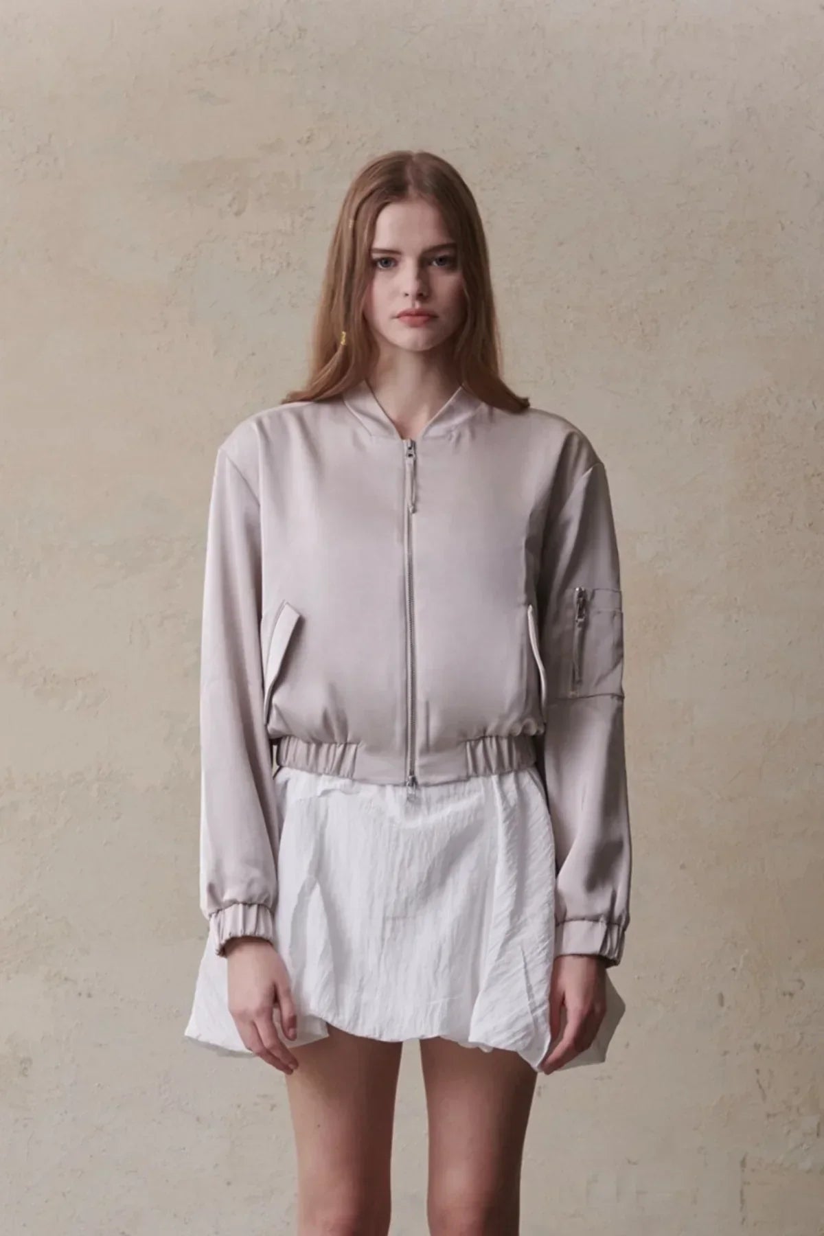 Front view of woman wearing beige satin bomber jacket and white skirt against textured wall