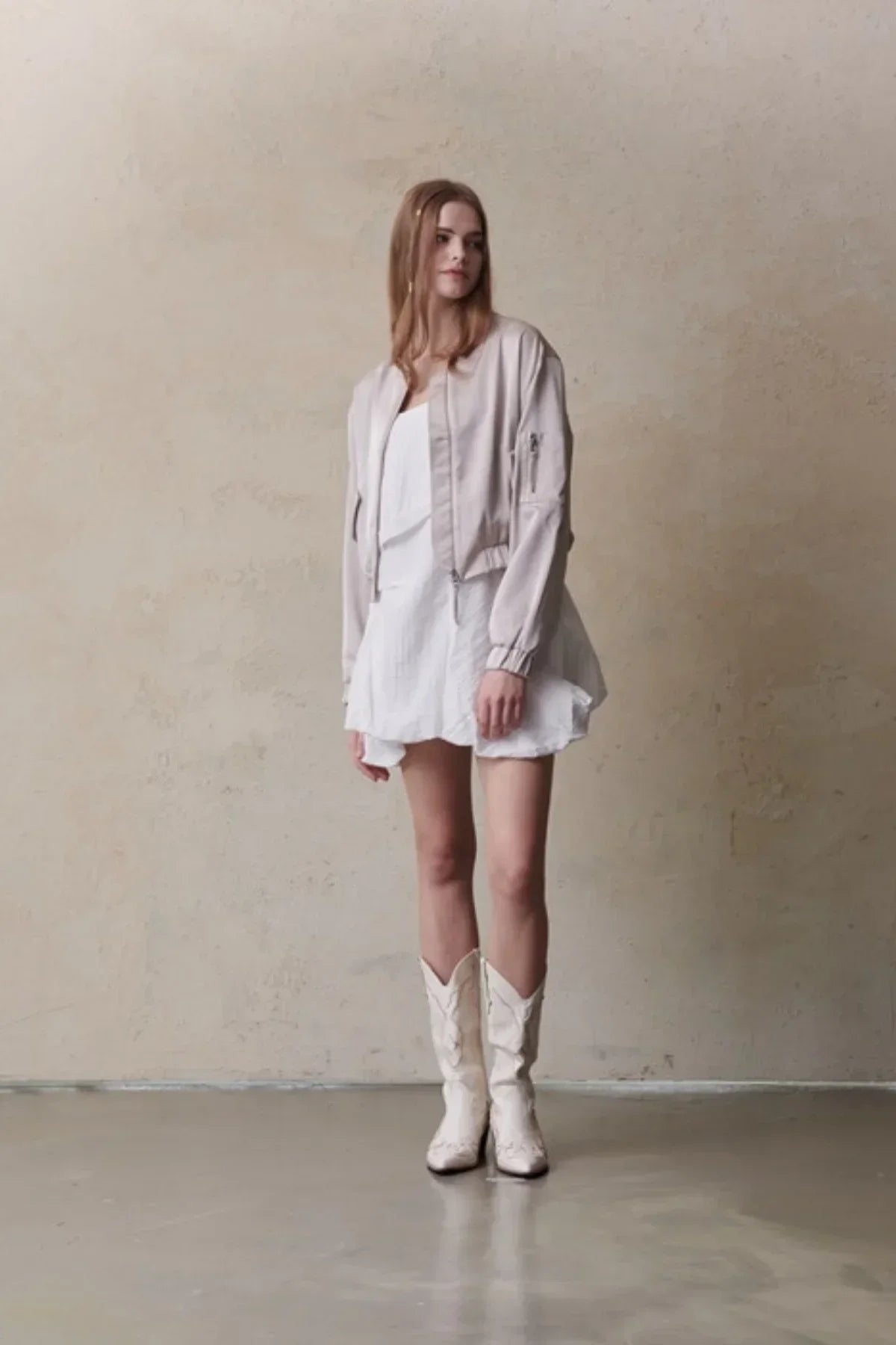 Model wearing beige satin bomber jacket, white mini dress, and white cowboy boots against neutral wall