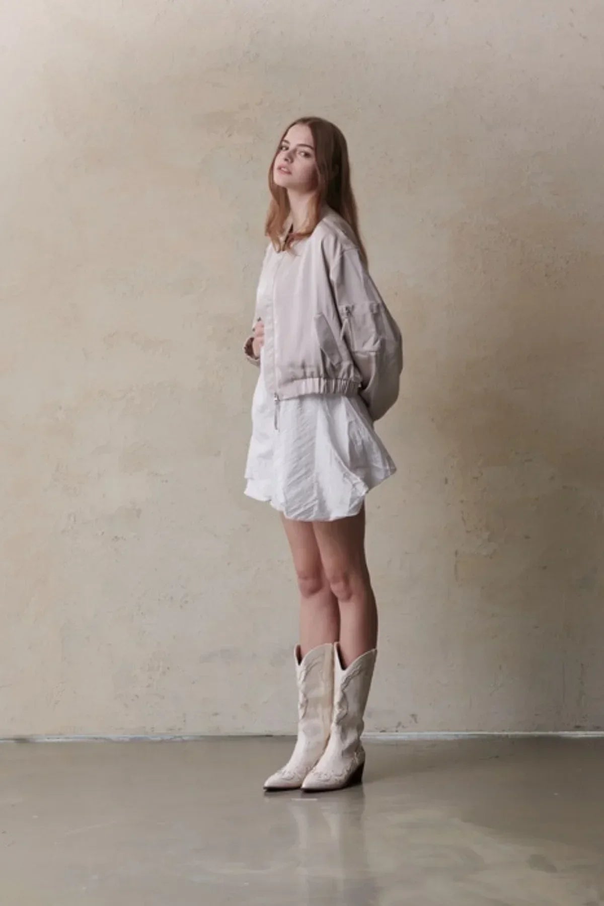 Model wearing beige satin bomber jacket, white dress, and cream cowboy boots against neutral wall