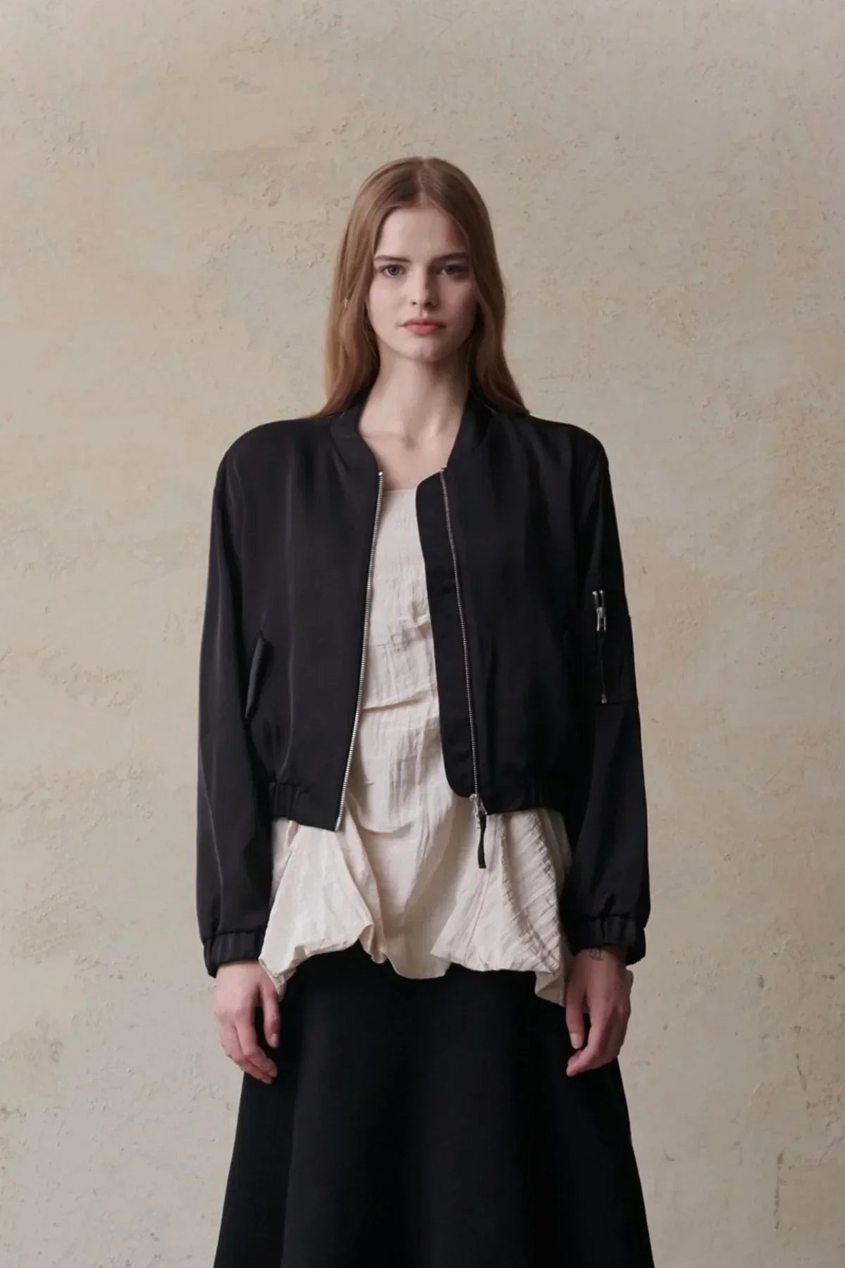 Model wearing a black crop satin bomber jacket with zipper and cream textured top against beige wall