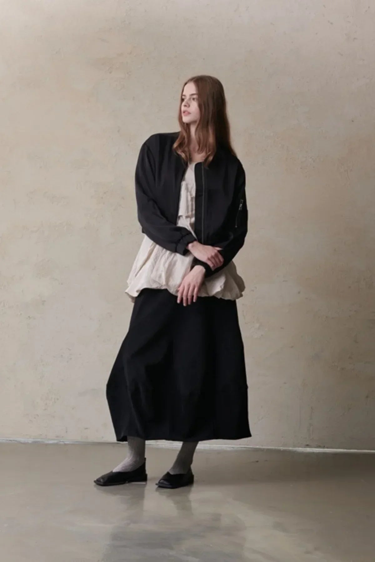 Model wearing black satin bomber jacket over beige top and black skirt, standing against textured wall