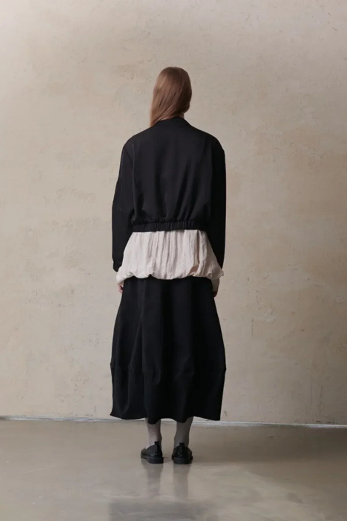 Model wearing black oversized skirt, white blouse, black cropped jacket, and black shoes in minimalist studio