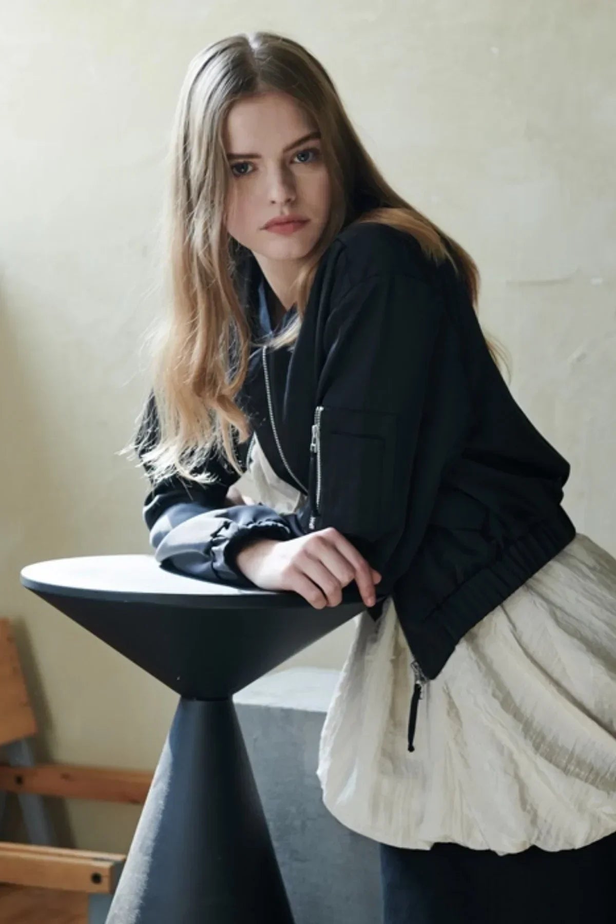 Young woman in black satin bomber jacket leaning on modern black cone-shaped table in minimal indoor setting