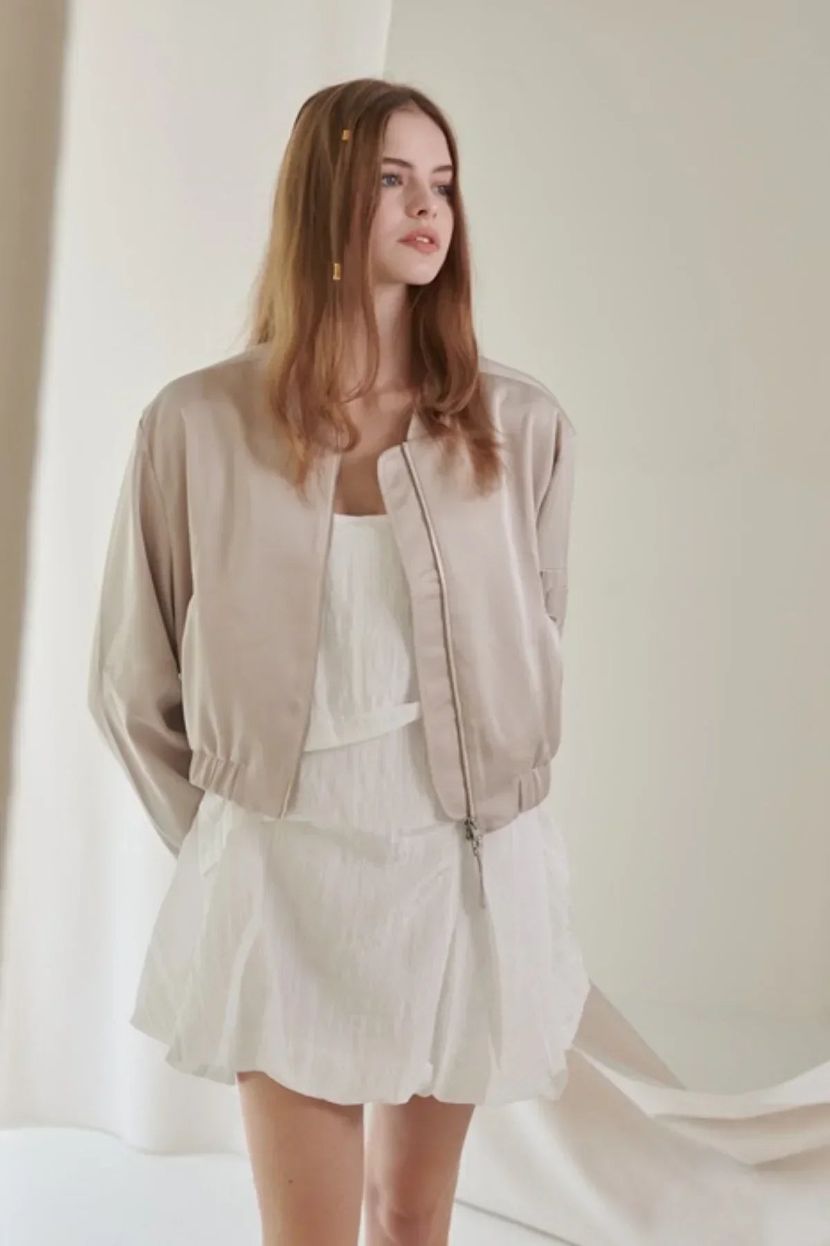 Young woman wearing a beige satin bomber jacket over a white dress in a minimalist indoor setting