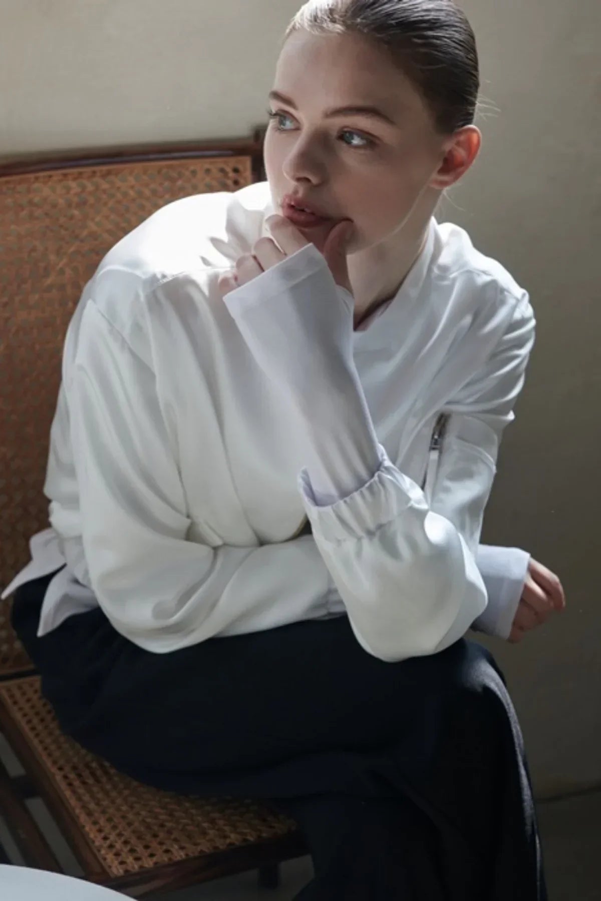 Woman wearing a white satin bomber jacket sitting on a woven chair in soft natural light
