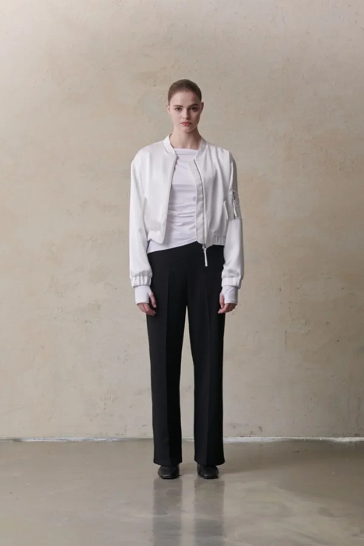 Model wearing a white satin bomber jacket, white top, black pants, and black shoes in a studio