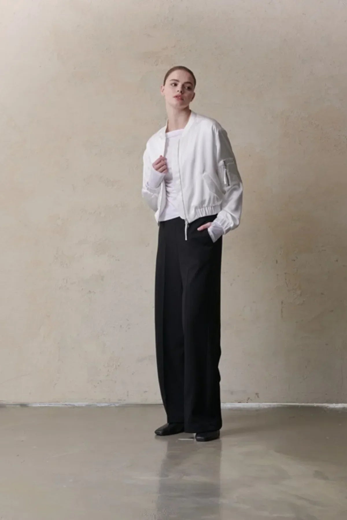 model wearing white satin bomber jacket with black wide-leg pants in minimalist indoor setting