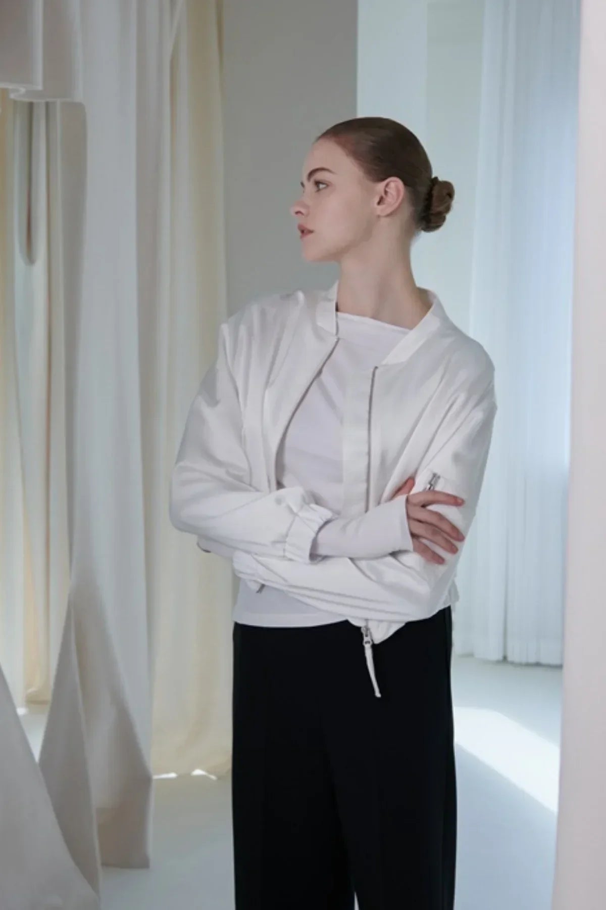 Young woman in a white satin bomber jacket and black pants standing in a bright, minimalist room