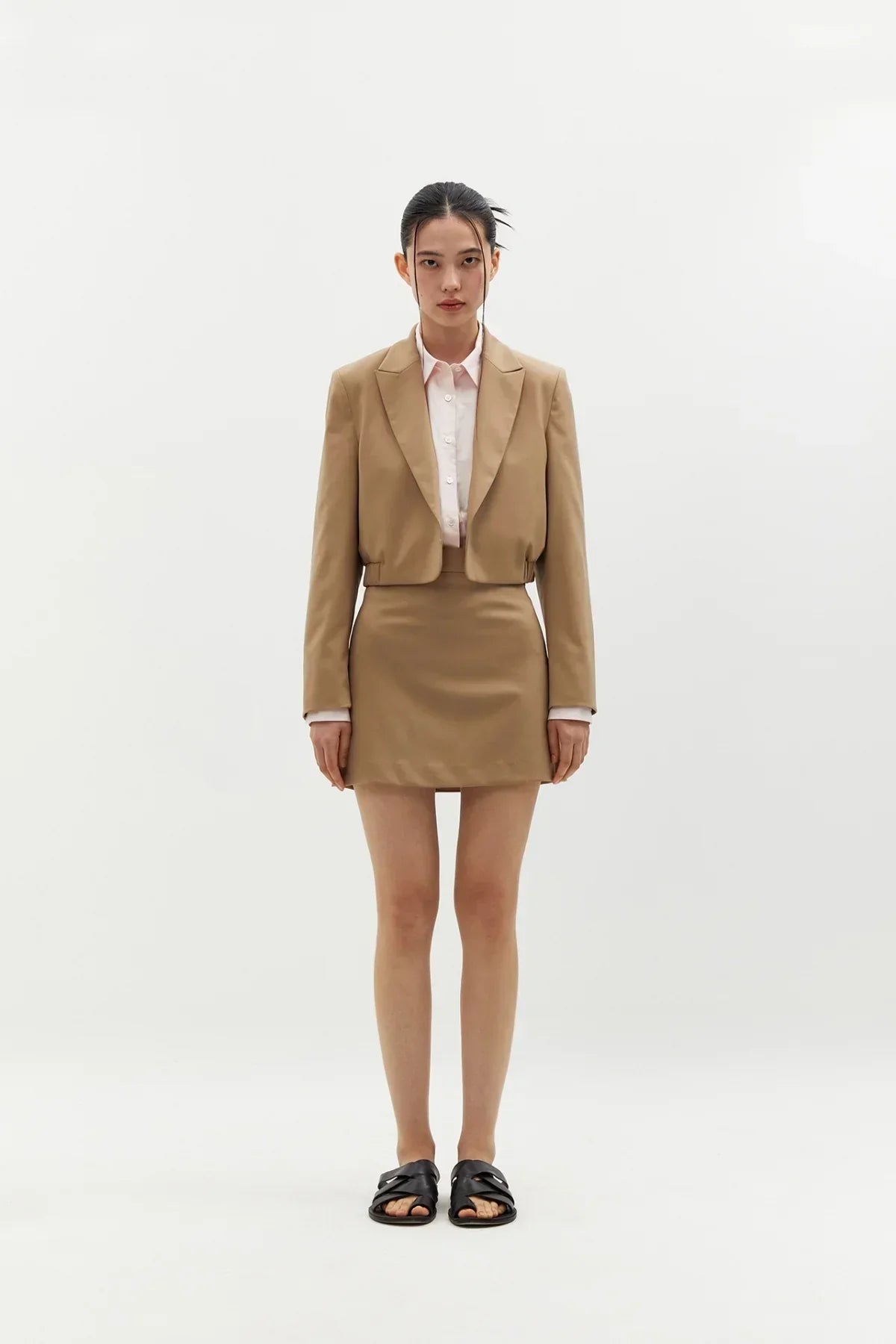 Woman wearing beige cropped blazer and matching mini skirt with pink shirt and black sandals on white background