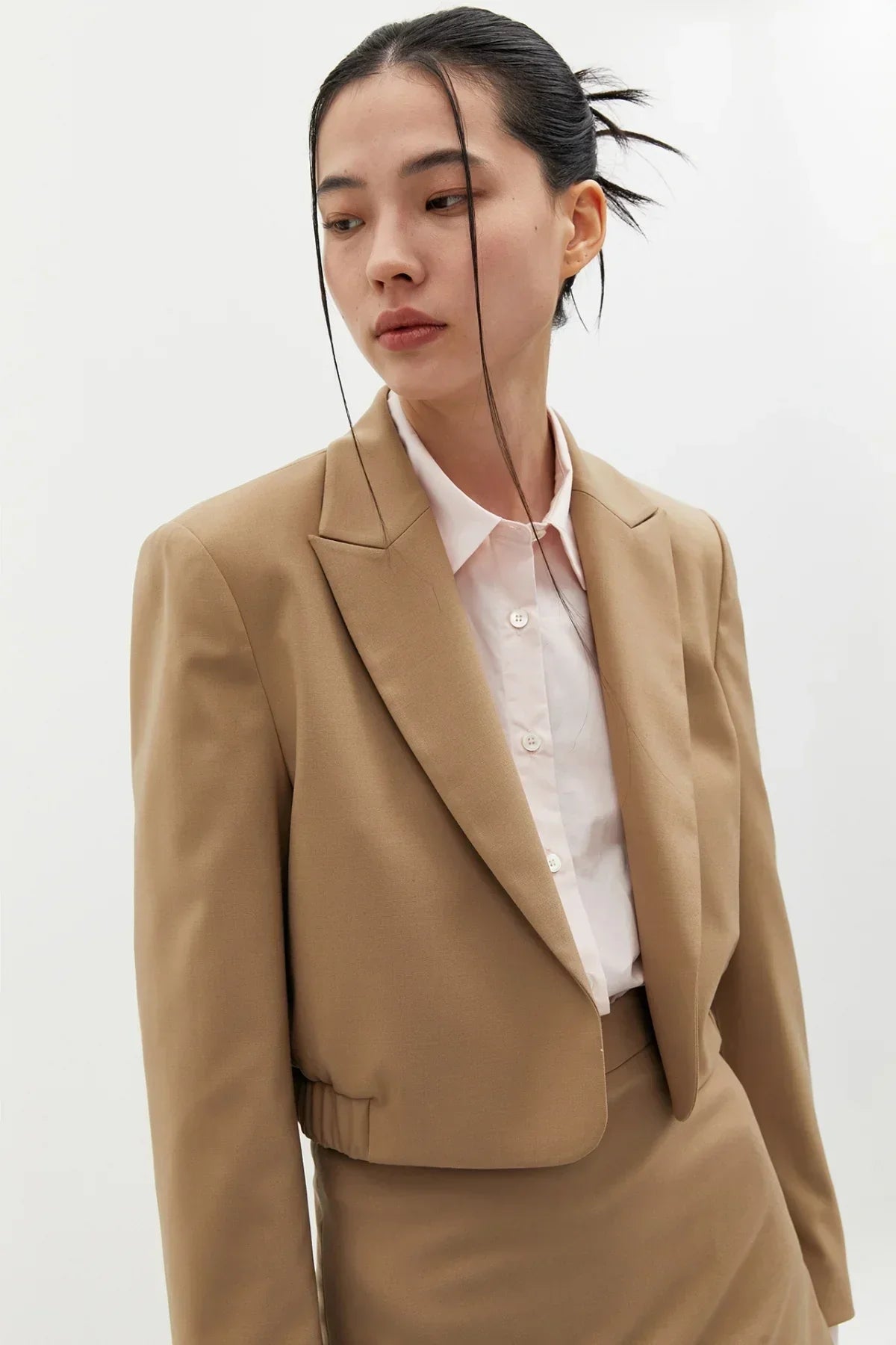 Woman wearing beige cropped wool blazer and matching skirt with white button-up shirt in studio